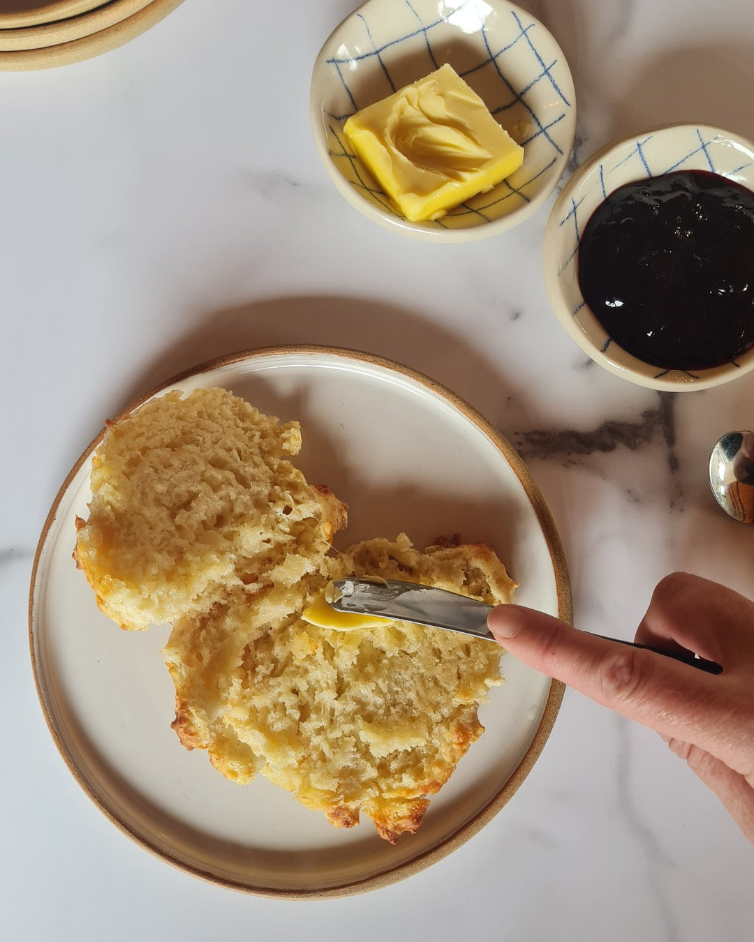 A hand holding a small knife is buttering a cheese scone sitting on a Meg Roulston Ceramcs white ceramic plate with a buff edge.  Two condiment bowls that have a white with blue check design sit above, one holds butter and one holds jam.