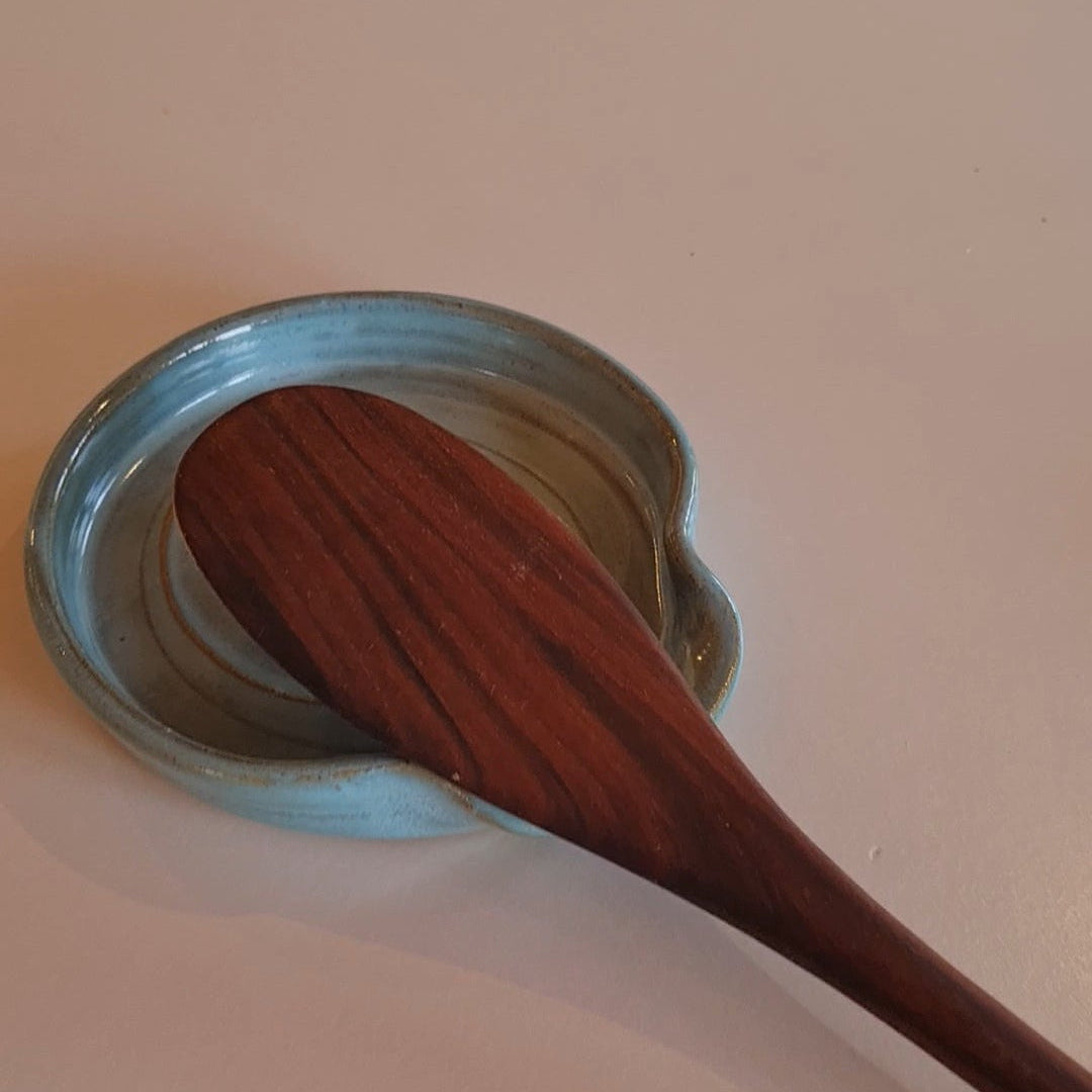 Rustic blue ceramic spoon rest with wooden spoon resting in it .