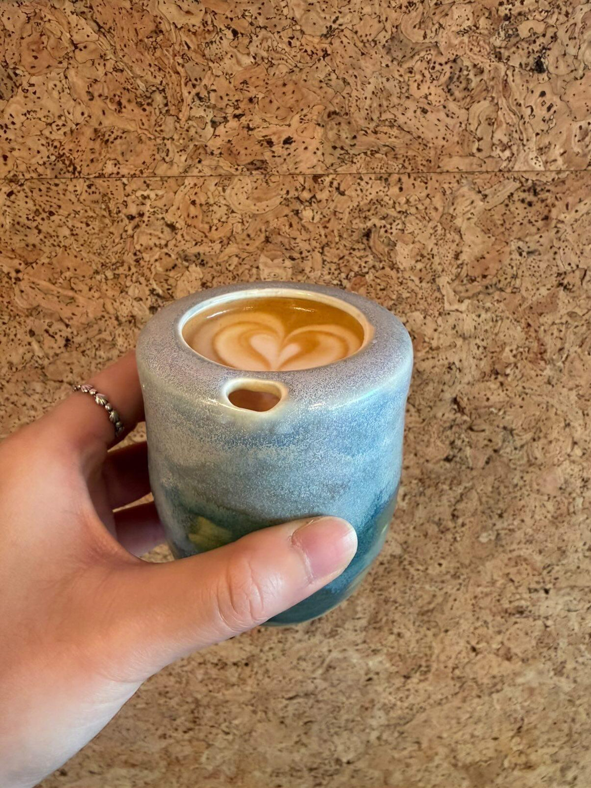 Coastal glaze ceramic takeaway tumbler held in hand with latte art, cork wall backdrop