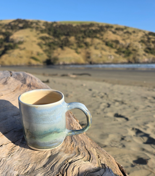 Ceramic mug glazed in a coastal landscape design sitting on driftwood in Okains Bay