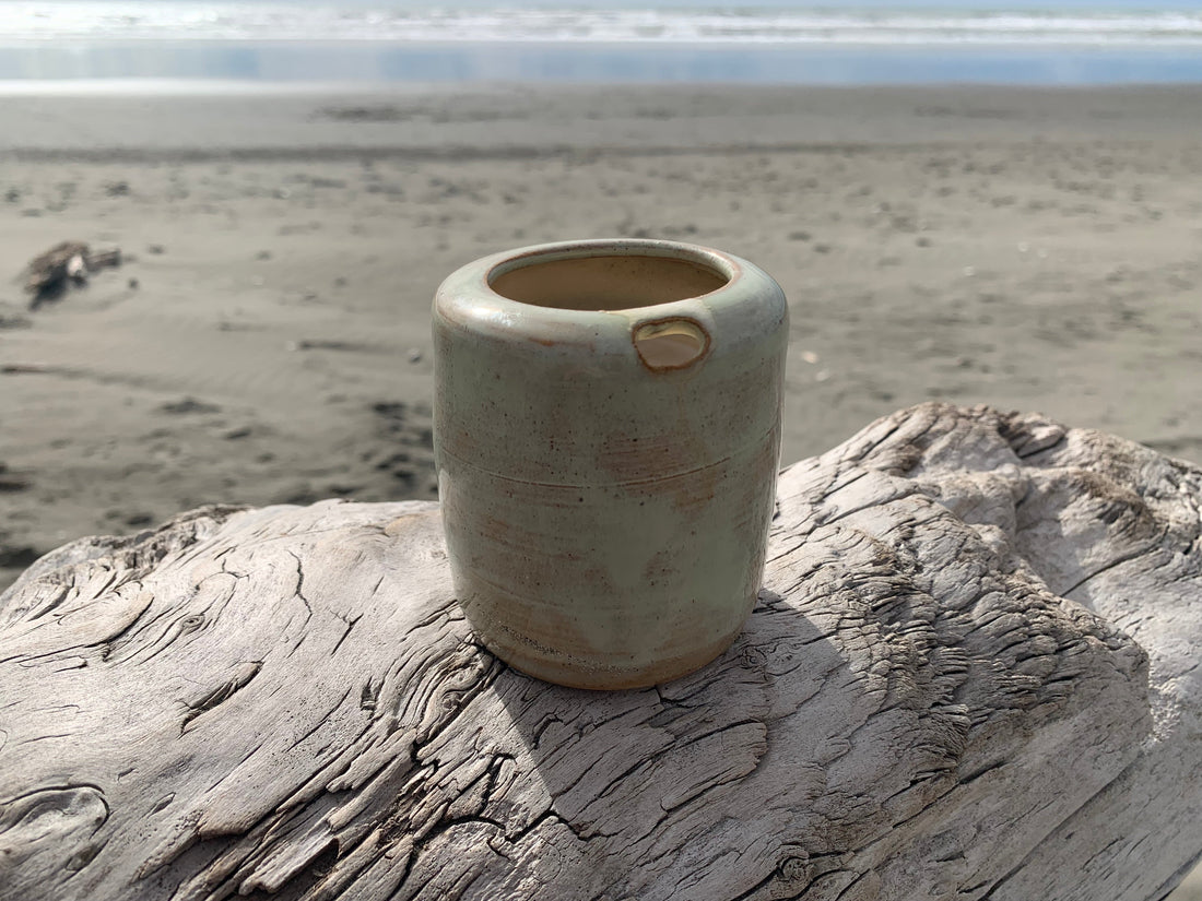 Takeaway tumbler by Meg Roulston Ceramics sits on driftwood in foreground on a beach with sea behind