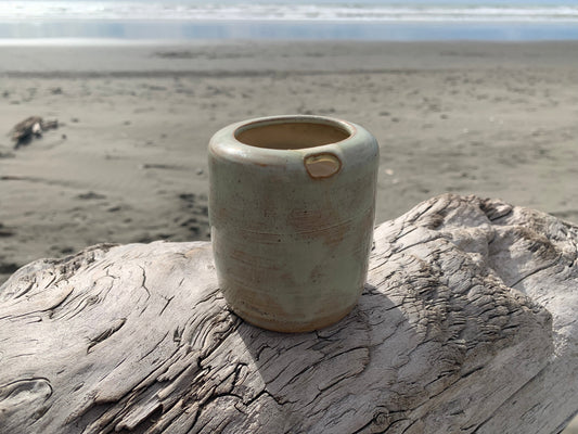 Takeaway tumbler by Meg Roulston Ceramics sits on driftwood in foreground on a beach with sea behind