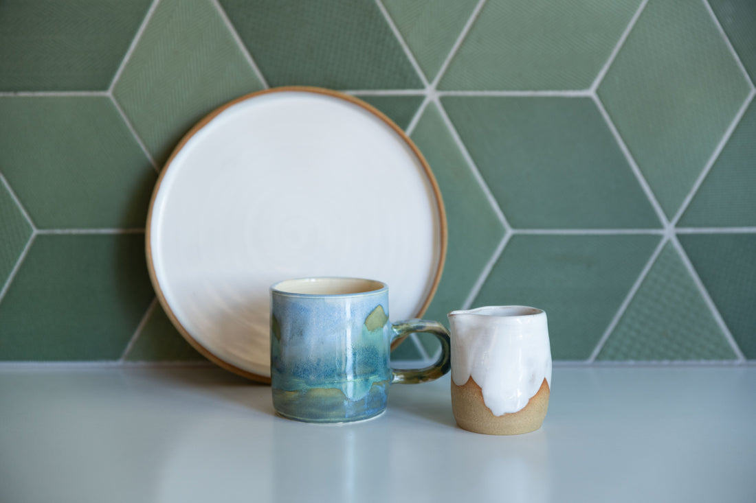 White plate with buff edge leans agains green tile splashback with a Coastal Landscape Mug and little white jug in front on white bench.  Meg Roulston Ceramics