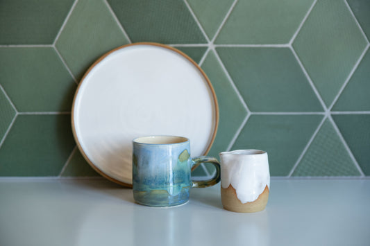 White plate with buff edge leans agains green tile splashback with a Coastal Landscape Mug and little white jug in front on white bench.  Meg Roulston Ceramics