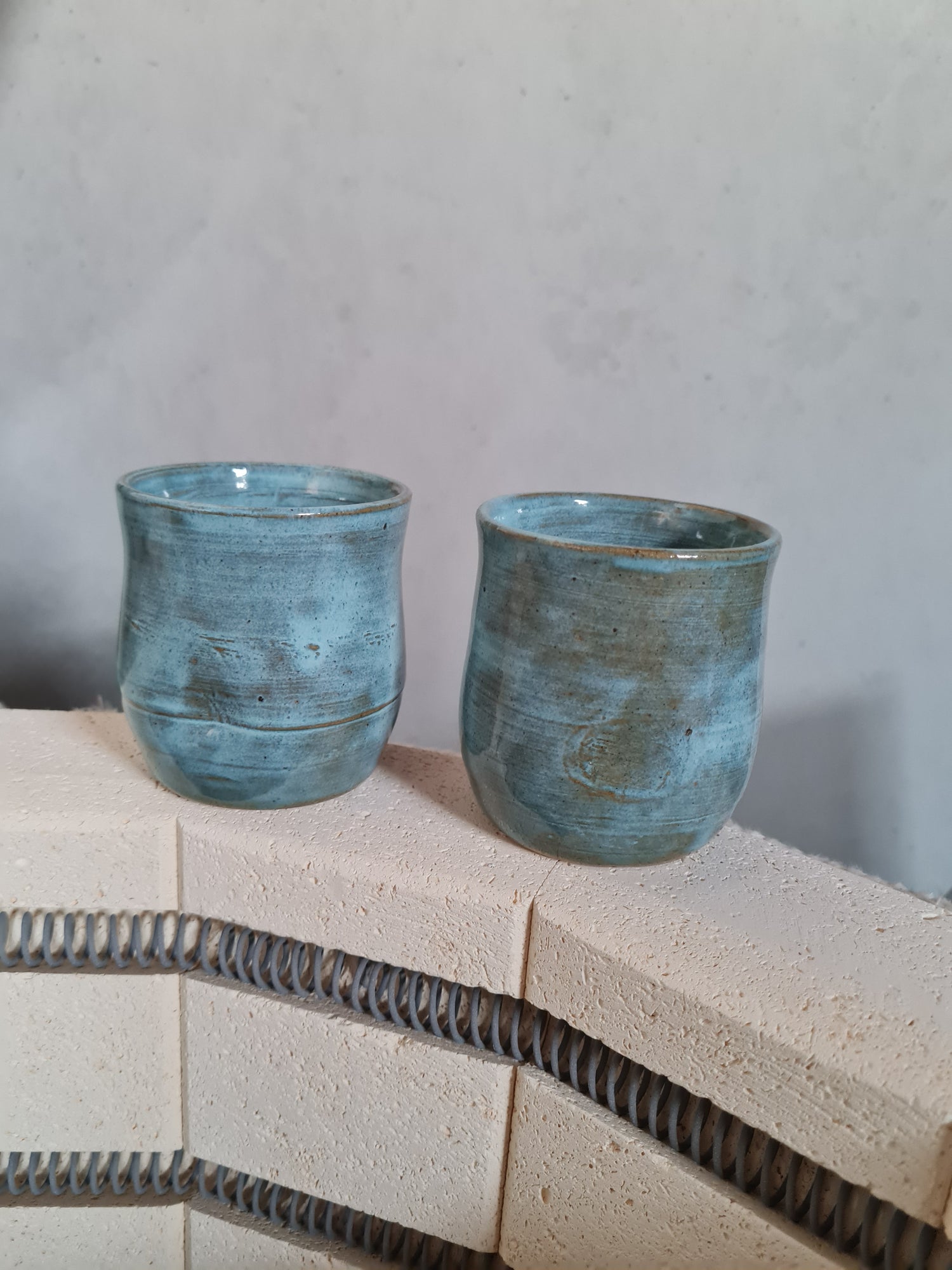 Two ceramic tumblers sitting on edge of an open kiln. The tumblers are a buff clay glazed in rustic blue. There is a comfort dimple on the tumbler to rest your thumb.