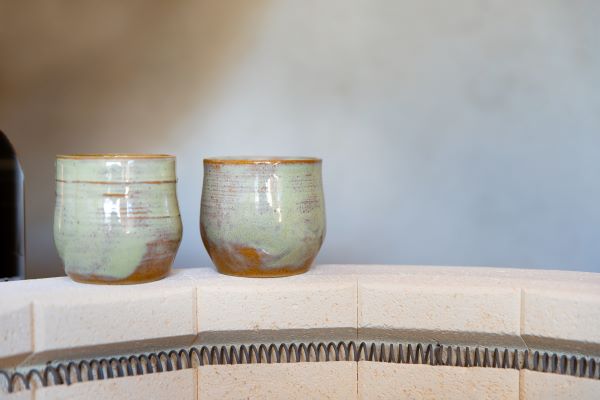 Two seagrass green ceramic tumblers with a belly shape sit on the edge of an open potters kiln.