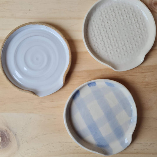 Three ceramic spoon rests, check, white daisy and whit with buff edge, sitting on a plywood backrground