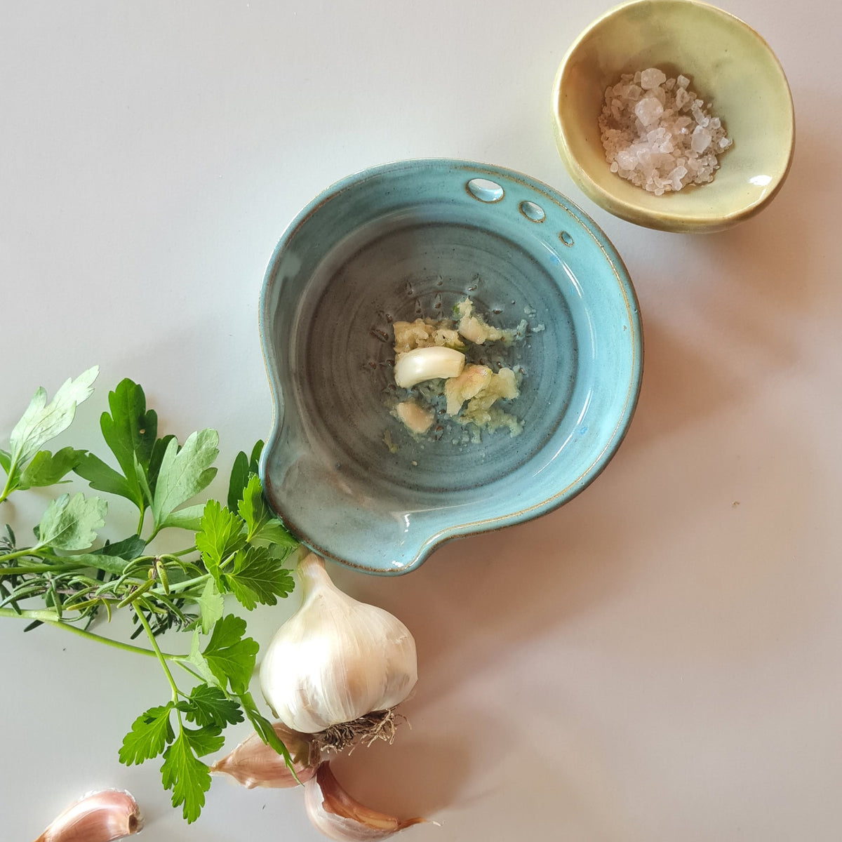 Rustic blue ceramic 3-in-1 garlic grater in use with freshly grated garlic, fresh herbs and a pinch bowl of salt 