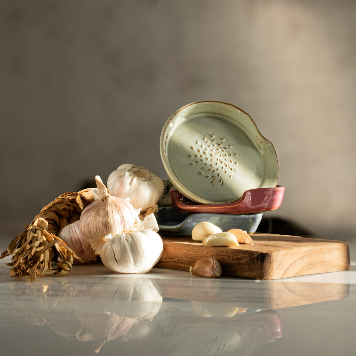 Three ceramic 3-in-1 garlic graters in seagrass green, pink and rustic blue stacked on a wooden board with garlic bulbs 