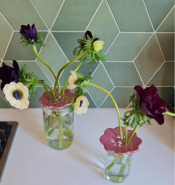 Two pink ceramic flower frogs holding anemones in glass jars on a kitchen bench, green tile backdrop 