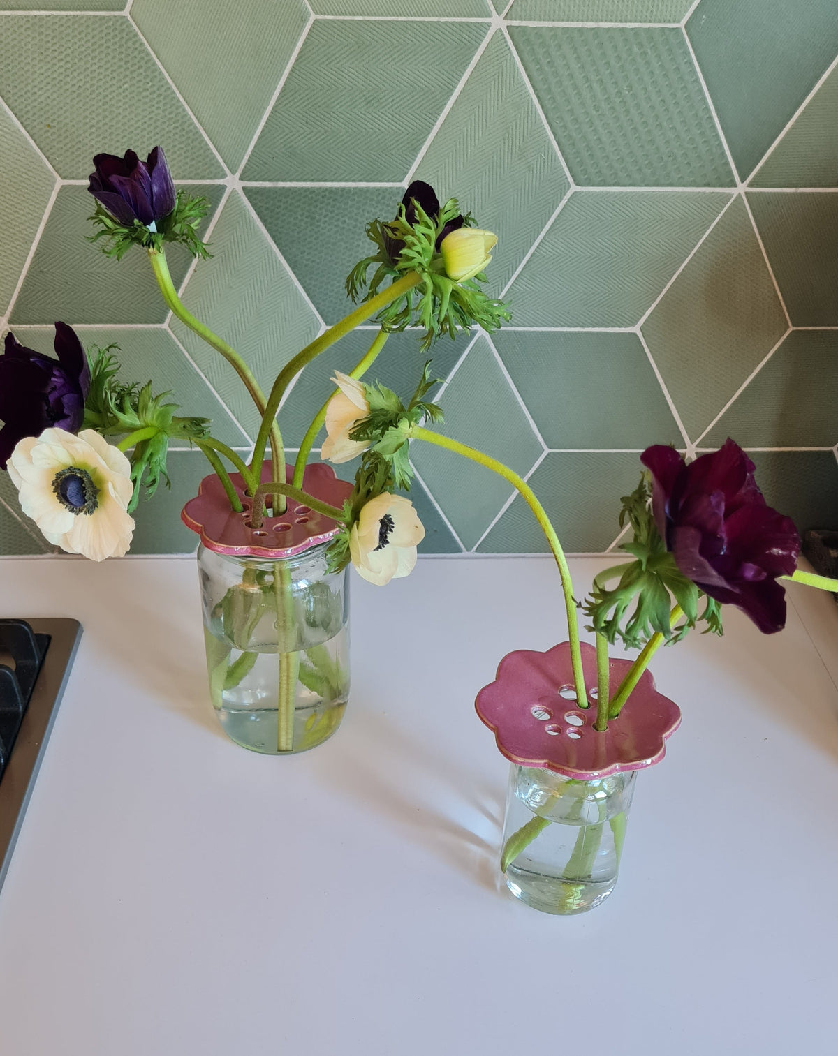 Two pink ceramic flower frogs holding anemones in glass jars on a kitchen bench, green tile backdrop