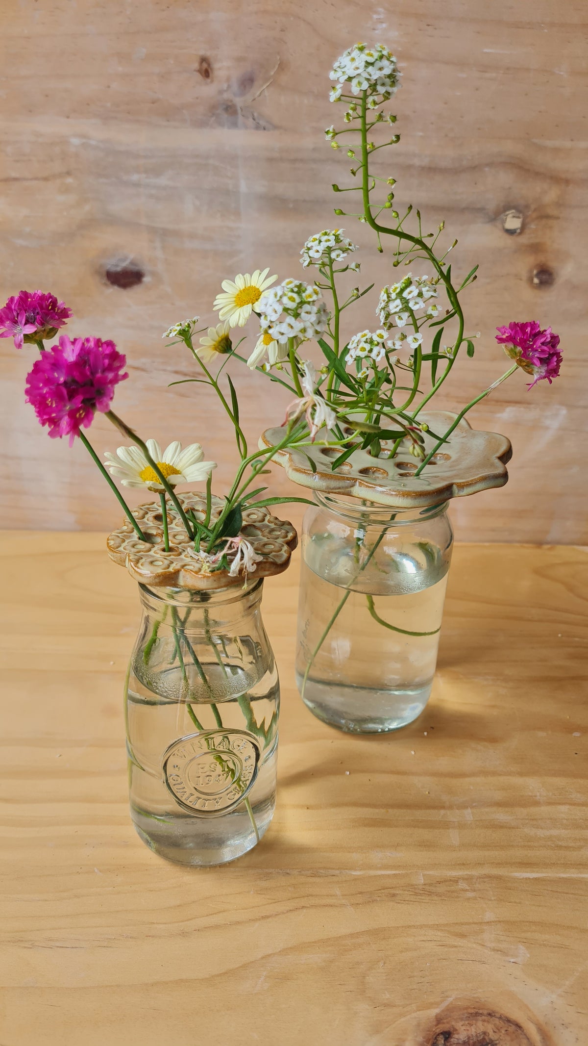 Two seagrass green ceramic flower frogs holding wildflowers in small glass jars on a wooden surface 