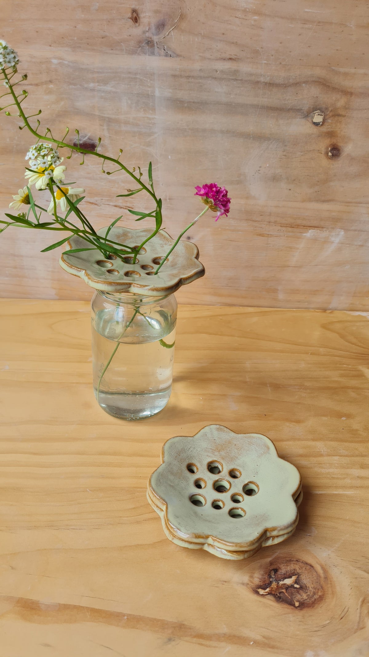Seagrass green ceramic flower frog holding wildflowers in a glass jar on a wooden surface, with two stacked frogs alongside 