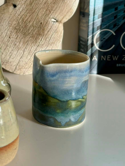 Handmade ceramic jug with coastal landscape glaze in blue and green, styled on a white shelf with driftwood 
