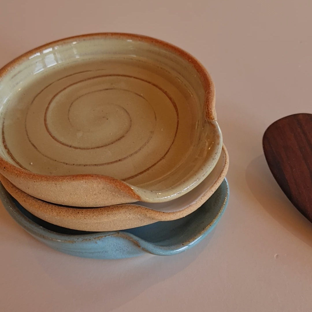 Stack of three ceramic spoon rests in white and rustic blue glazes with a wooden spoon, on a white surface 