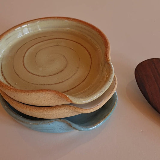 Stack of three ceramic spoon rests in white and rustic blue glazes with a wooden spoon, on a white surface 