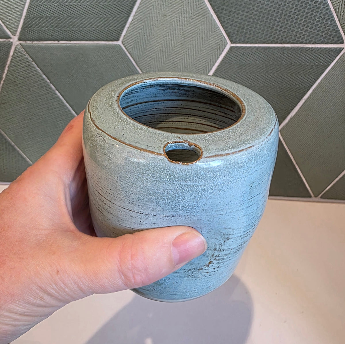 Rustic blue ceramic takeaway tumbler held in hand showing thumb notch and textured surface, green tile backdrop