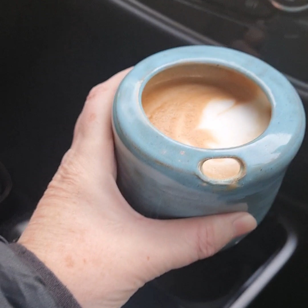 Rustic blue ceramic takeaway tumbler held in hand with a latte, in use outdoors 