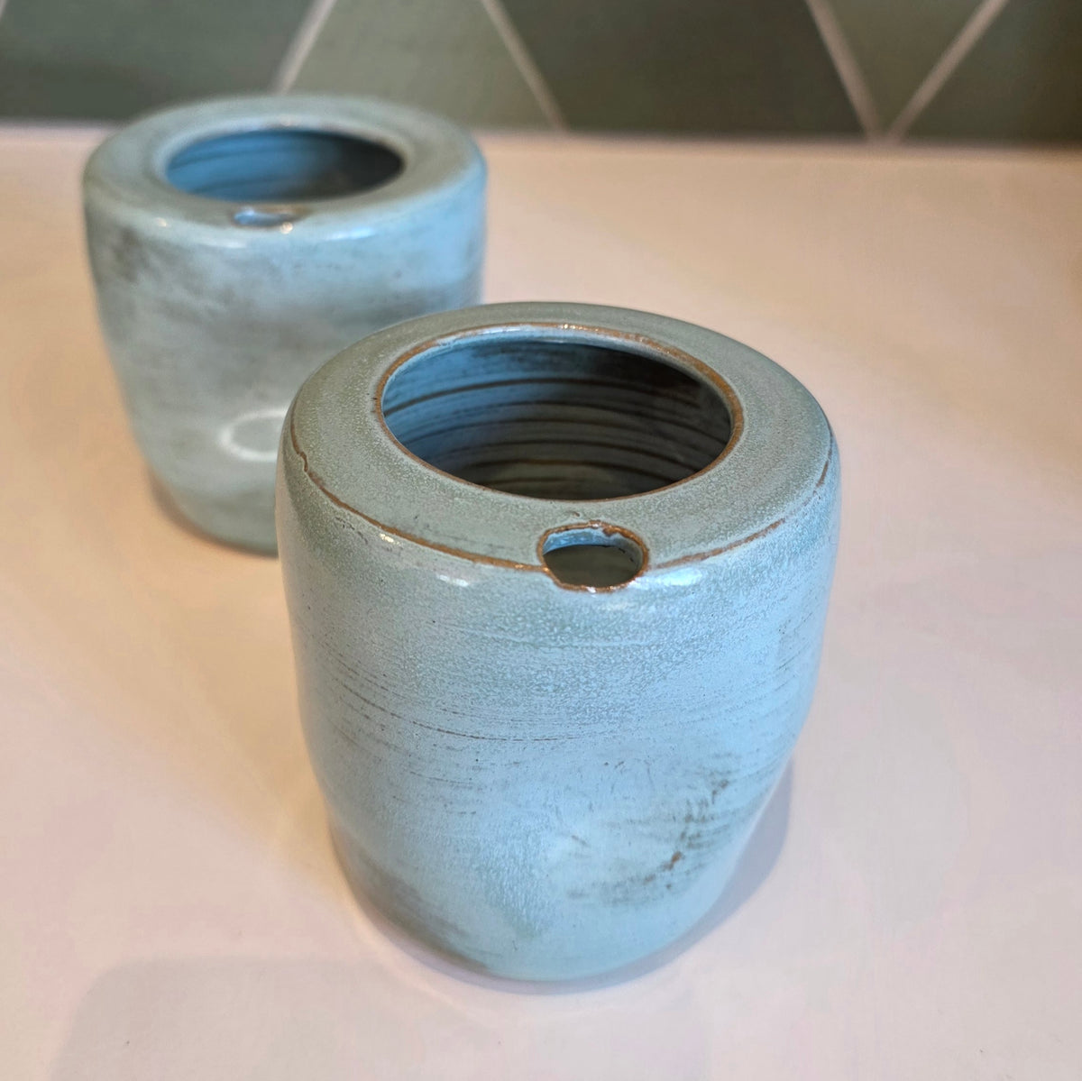Two rustic blue ceramic takeaway tumblers on a bench showing thumb notch detail, green tile backdrop 