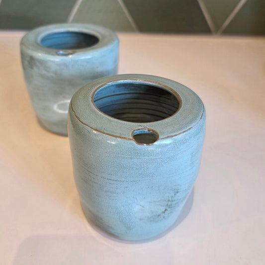 Two rustic blue ceramic takeaway tumblers on a bench showing thumb notch detail, green tile backdrop 