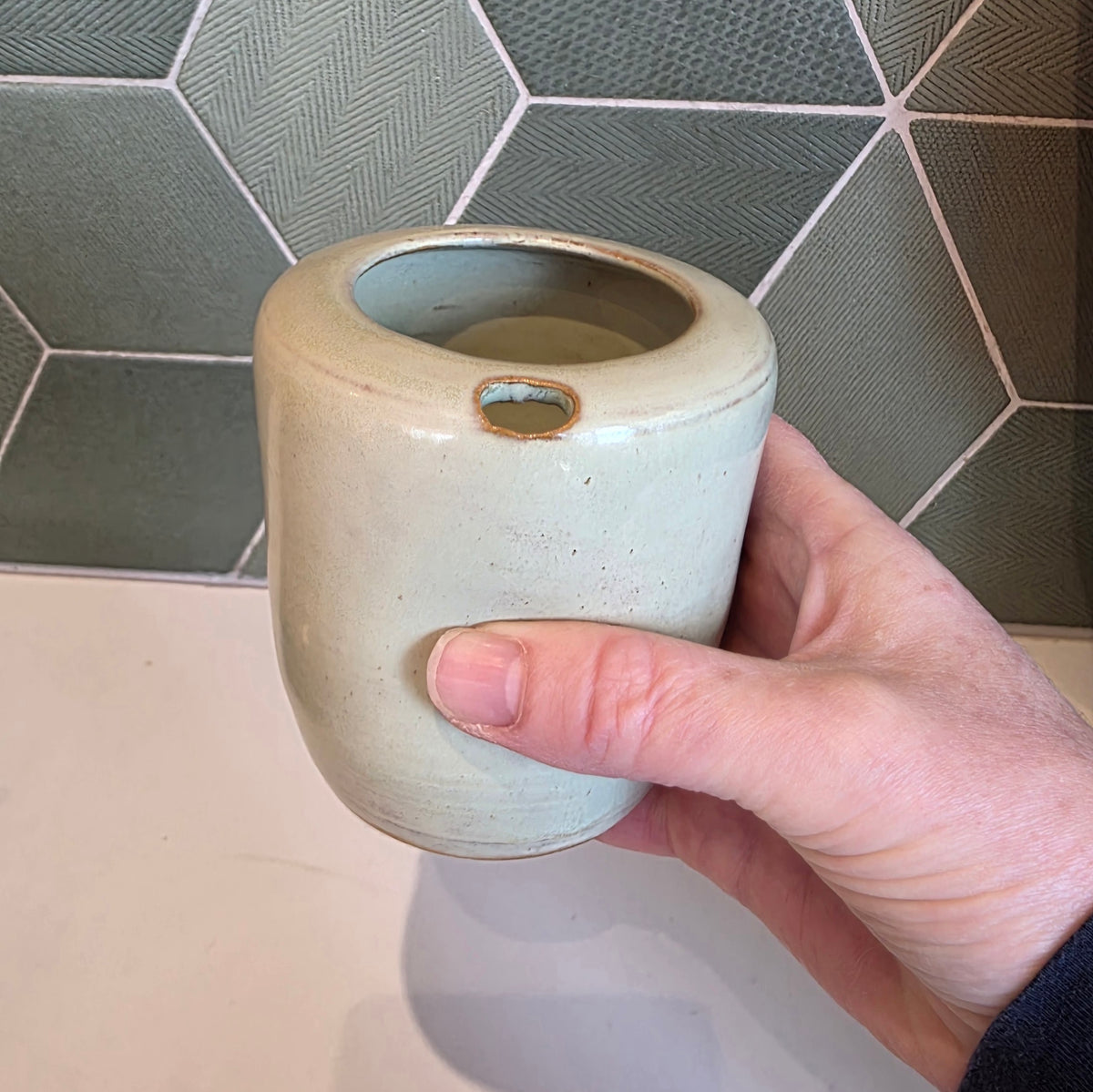 Seagrass green ceramic takeaway tumbler held in hand showing thumb notch and size, green tile backdrop 