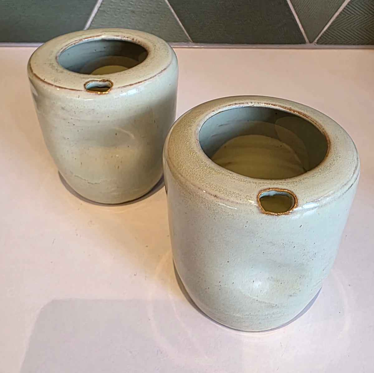 Two  seagrass green ceramic takeaway tumblers on a bench showing thumb notch detail, green tile backdrop