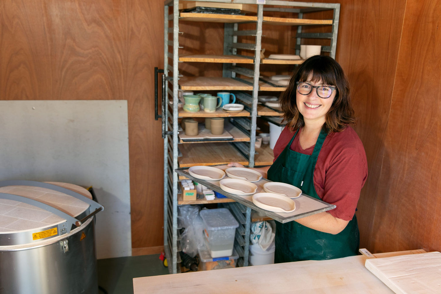 Potter Meg Roulston in her studio holding a wareboard full of spoon rests, she stands in front of a bakers trolly full of ceramics and her kiln is off to the right.