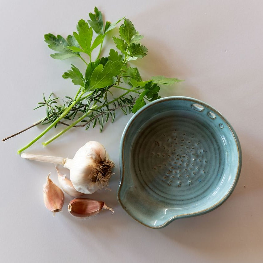 Flatlay rustic blue 3in1 grater spoon rest and herb stripper with bulb of garlic and bunch of herbs beside it
